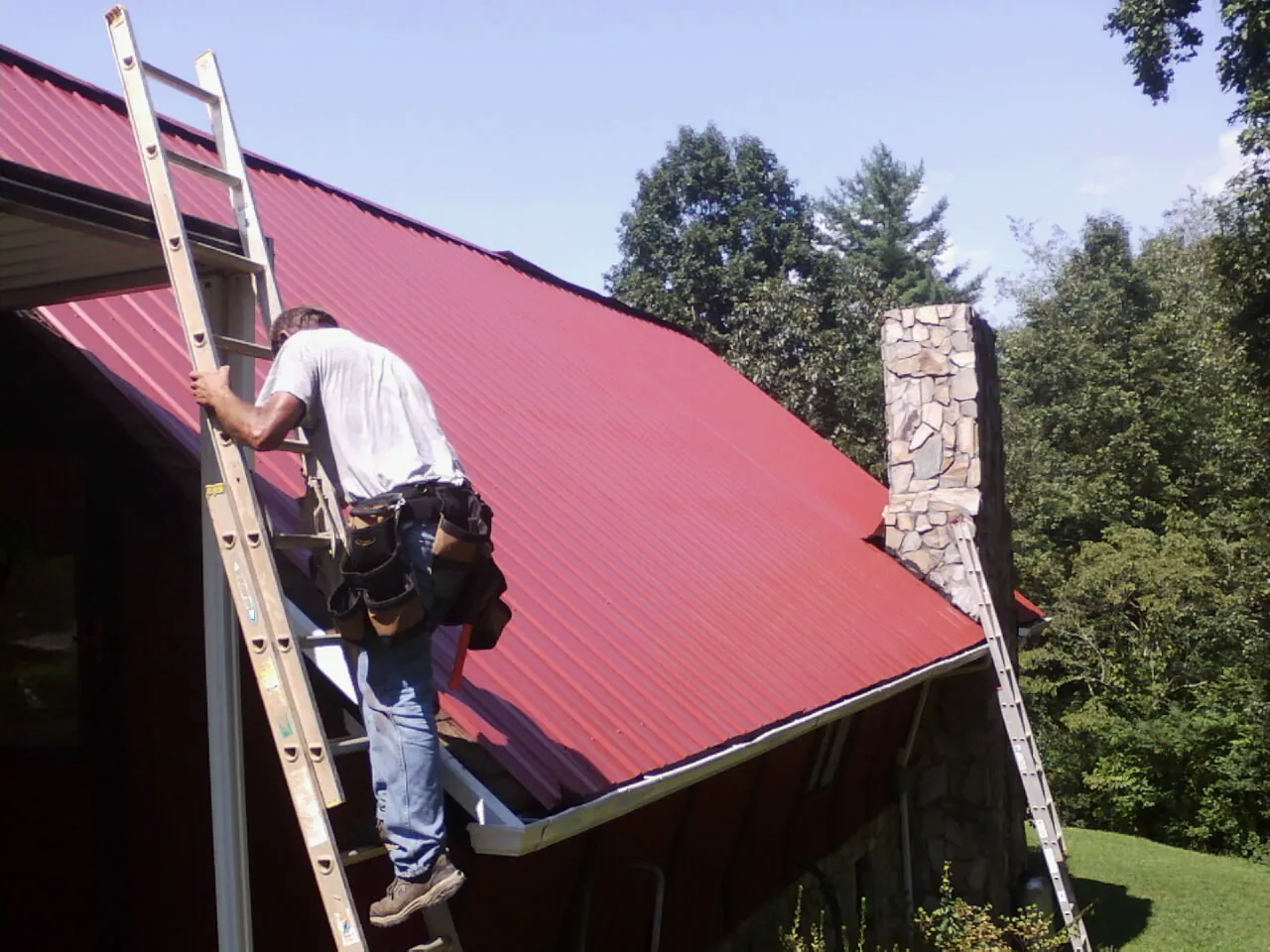 Roofer on a ladder beside a red metal roof and stone chimney — Soffit & Fascia Repair by Roofing Brothers in South Berwick